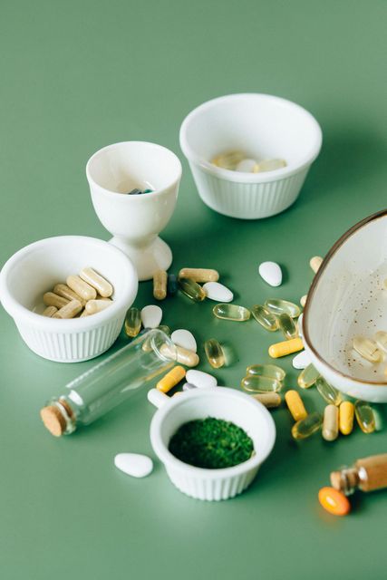 Assorted pills and capsules spilling from white bowls and glass bottles on a green surface.