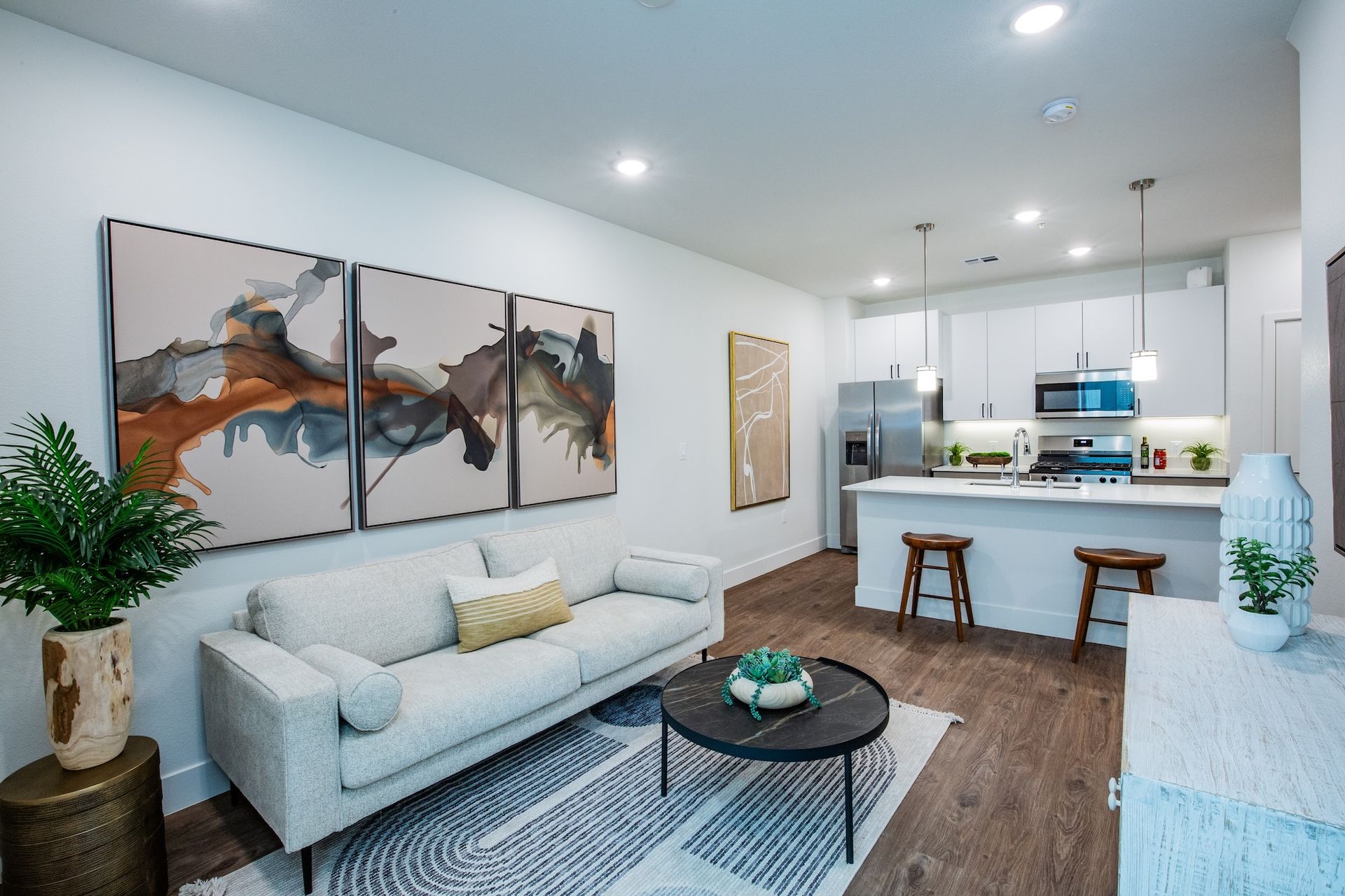 Modern apartment living room with open kitchen, white walls, abstract art, and wooden floors.