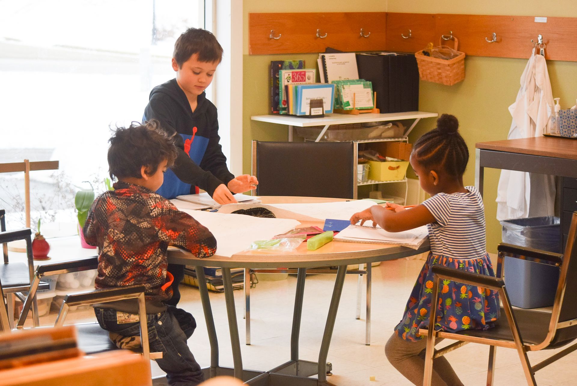 Children at a round table, drawing and working on a craft project in a classroom.