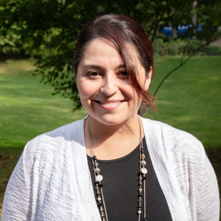 Woman with brown hair and a white cardigan smiles in a grassy outdoor setting.