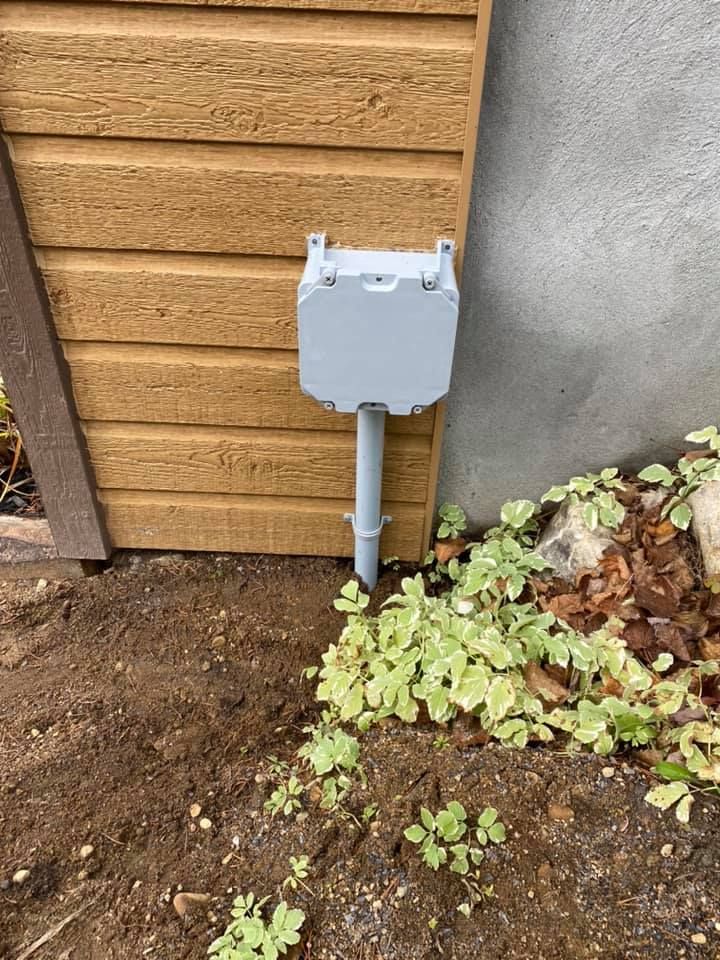 Gray utility box mounted on a pole, against a wooden wall and concrete, in a garden.