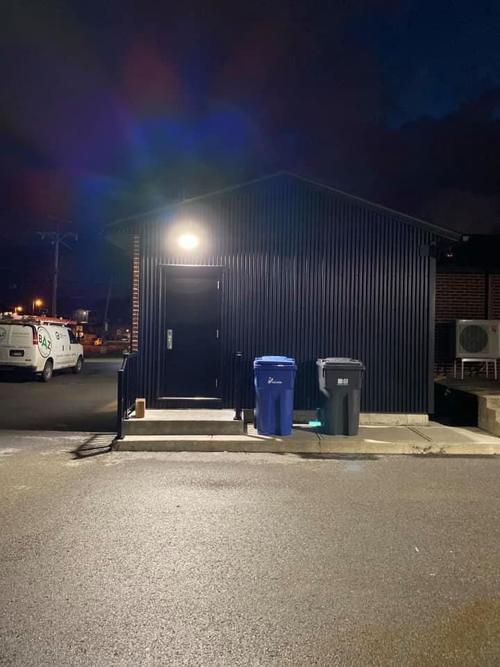 Dark corrugated metal building with open doorway illuminated by a light. Two trash bins in front.