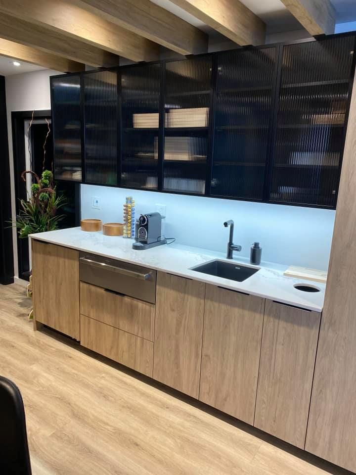 Modern kitchen with light wood cabinets, black-framed glass upper cabinets, stainless steel appliances, and a black sink.