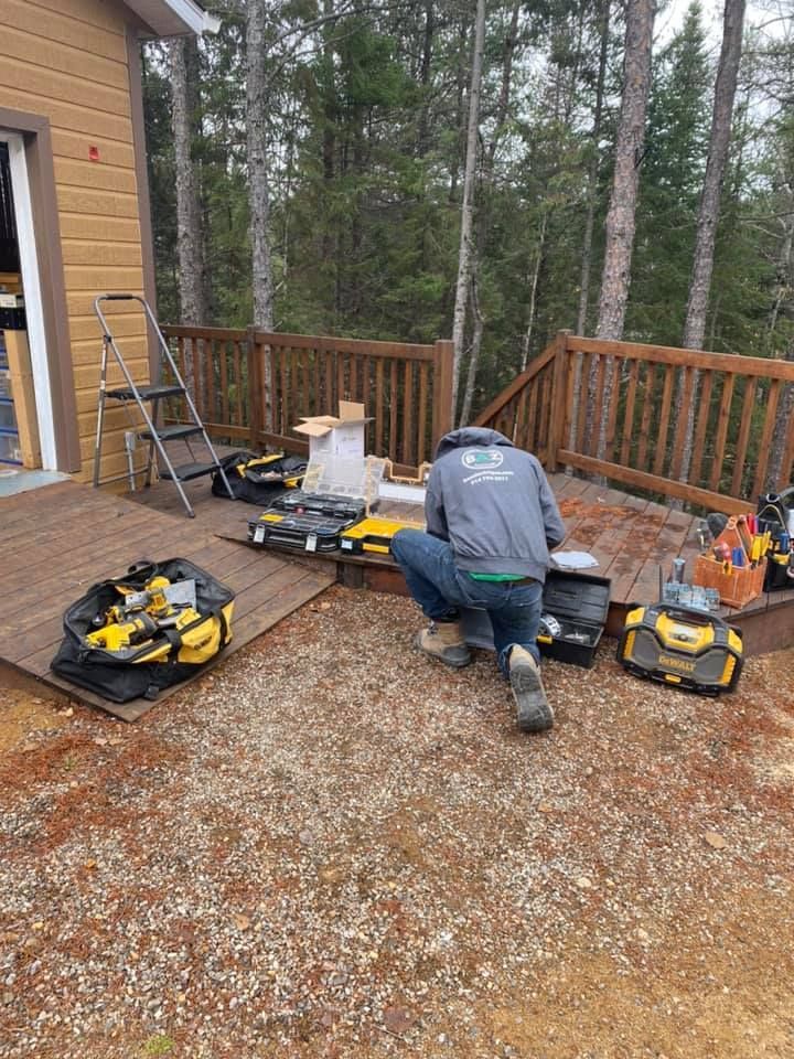 Person kneeling, sorting tools on a deck, with a building and woods in the background.