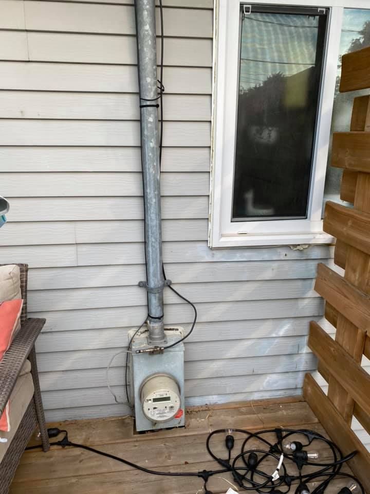 Electrical meter box mounted on a gray-sided house with a window and wooden deck.