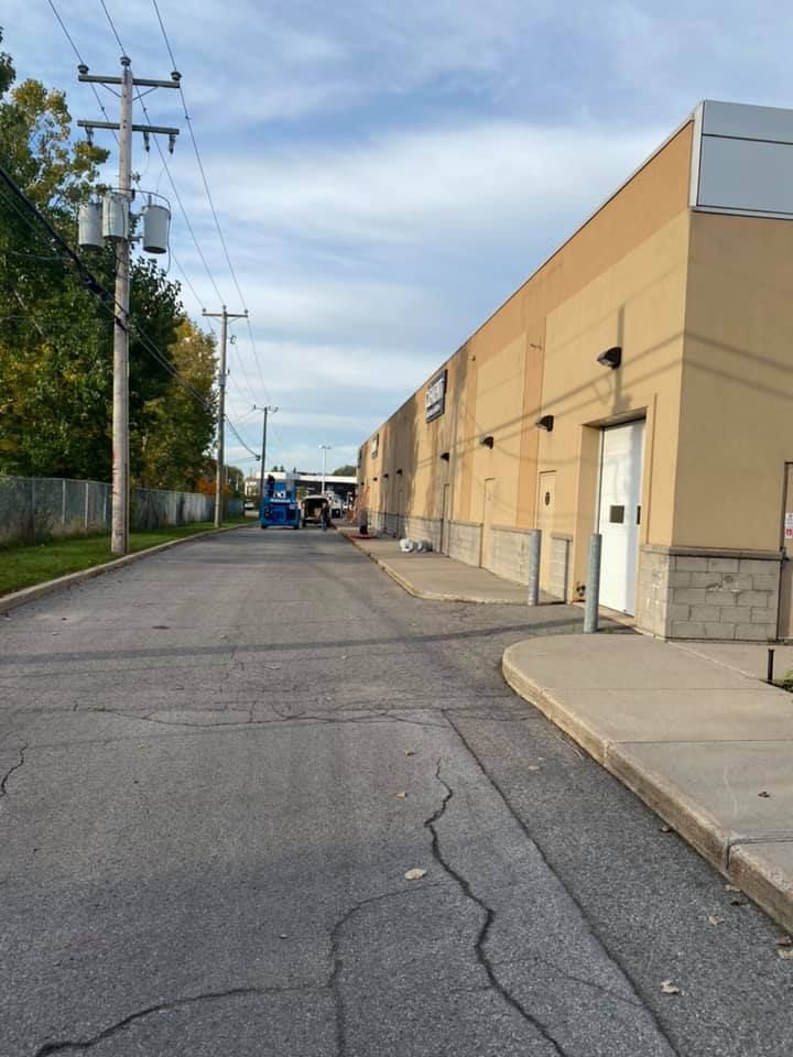 Alleyway beside tan industrial building with a white door. Blue truck in the distance under overcast sky.