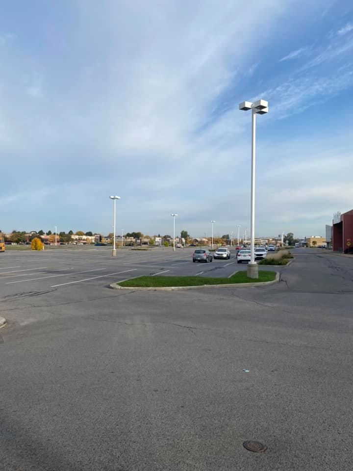 Empty asphalt parking lot with multiple tall white light poles and a few parked cars under a blue cloudy sky.