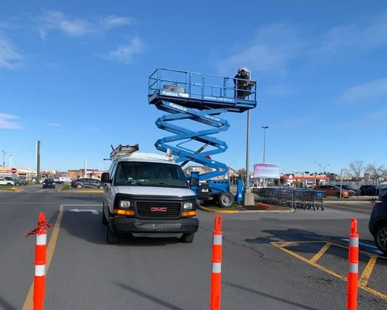 White van in parking lot, orange safety cones, blue scissor lift with person working, other vehicles and stores visible.