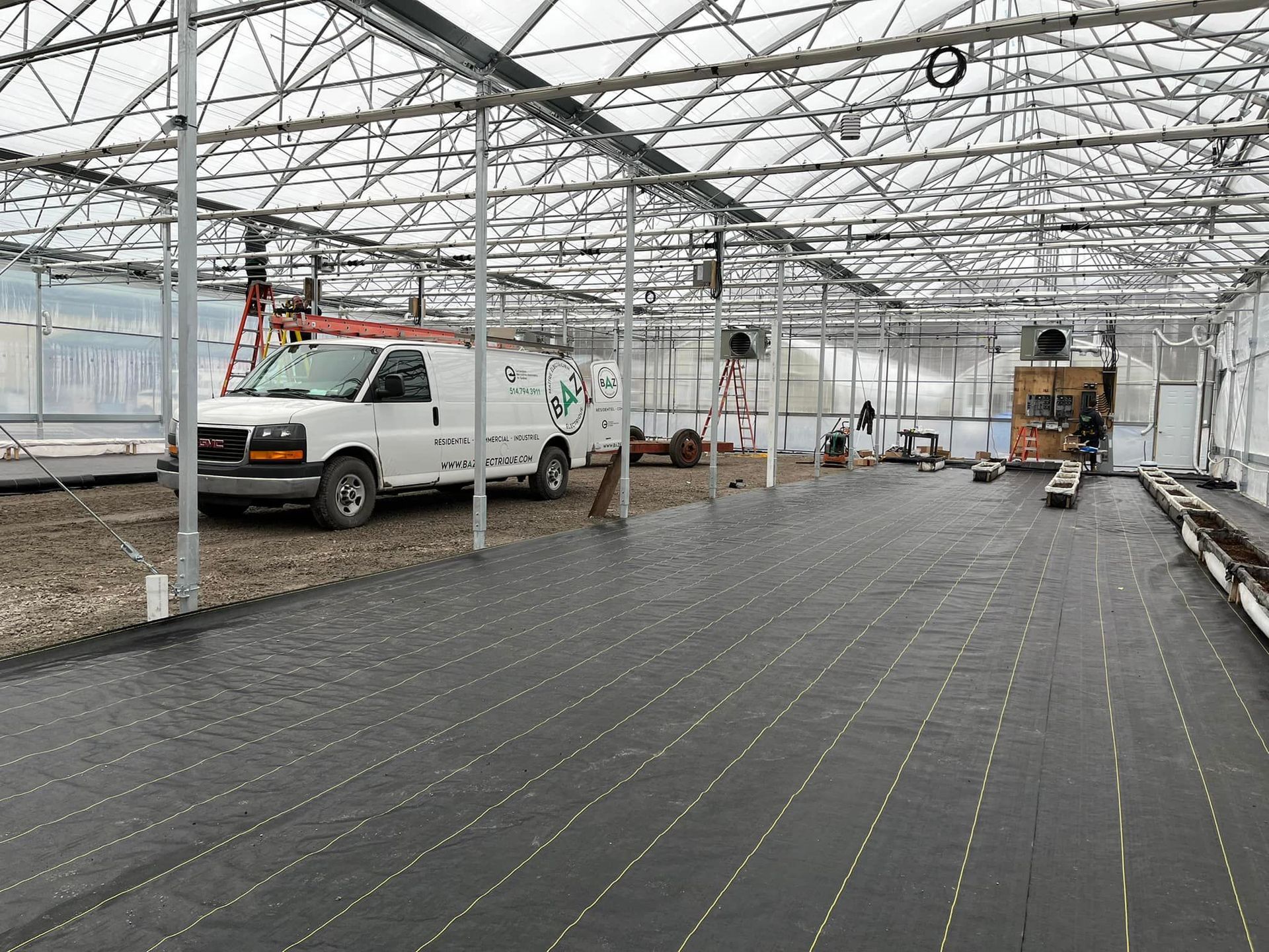Inside a greenhouse: white van, workers, and equipment on gravel floor with black weed barrier.