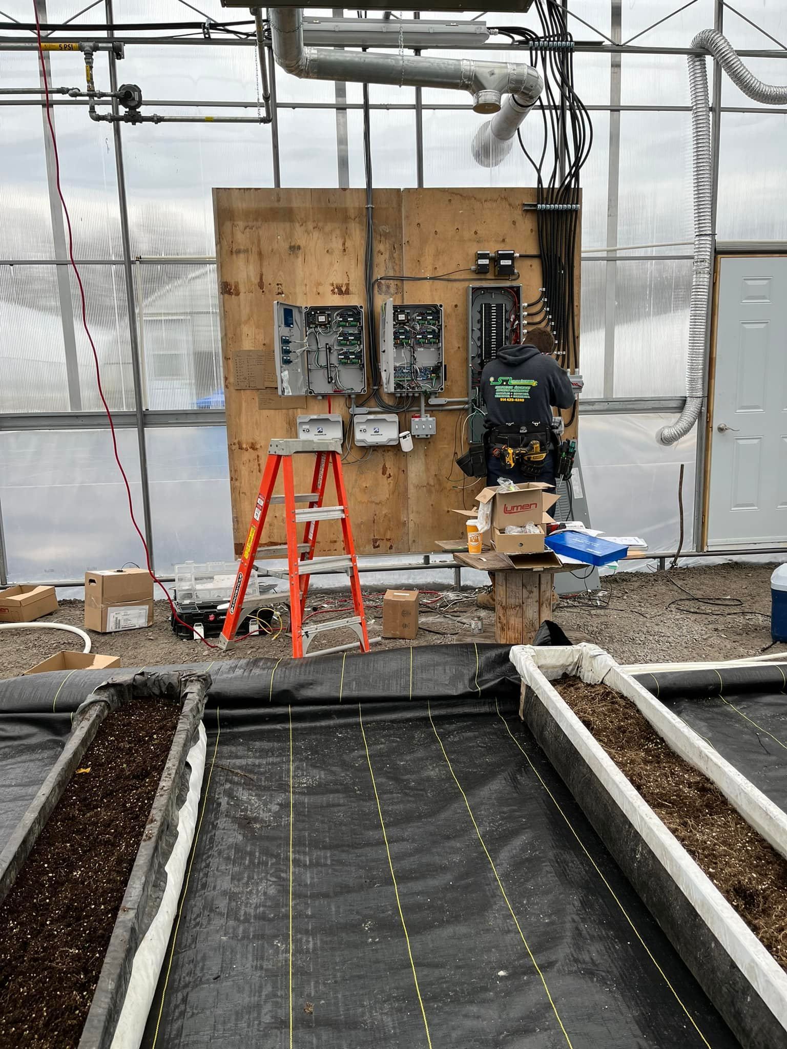 A person works on electrical panels inside a greenhouse. Electrical equipment is mounted on a wooden wall.