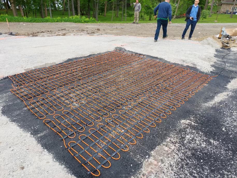 Installation of heated pavement, copper tubing grid laid on gravel; three men observe.