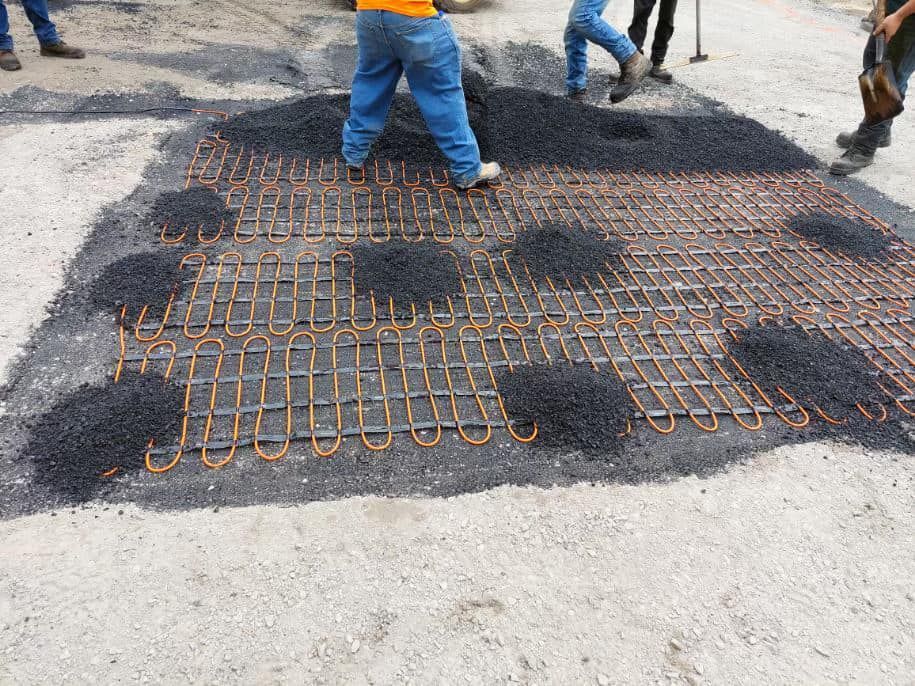 Workers laying asphalt over a metal grid on a road. Dark asphalt, blue pants, orange shirt.