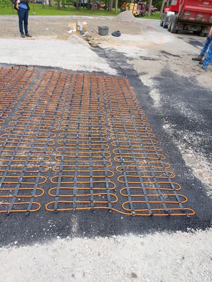 Copper tubing grid laid on asphalt for radiant heating. Construction site with truck and workers.