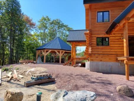 Log cabin with a gazebo under construction; gravel yard, trees in background, sunny day.