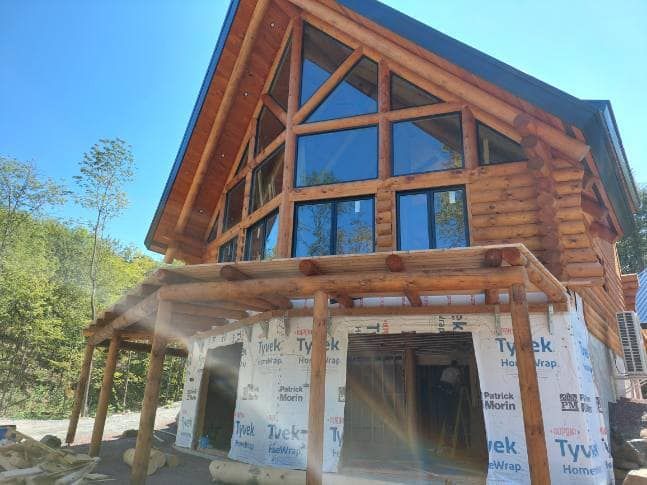 Log cabin under construction with large windows and a covered porch.