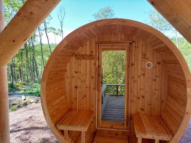 Wooden barrel sauna interior with benches, open doorway to forest view.