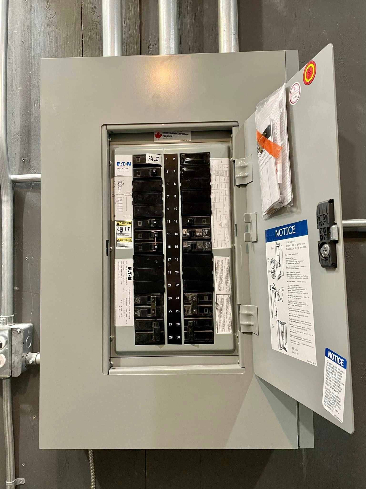 Electrical panel with open door, showing circuit breakers and labels. Gray metal box on wall, pipes nearby.