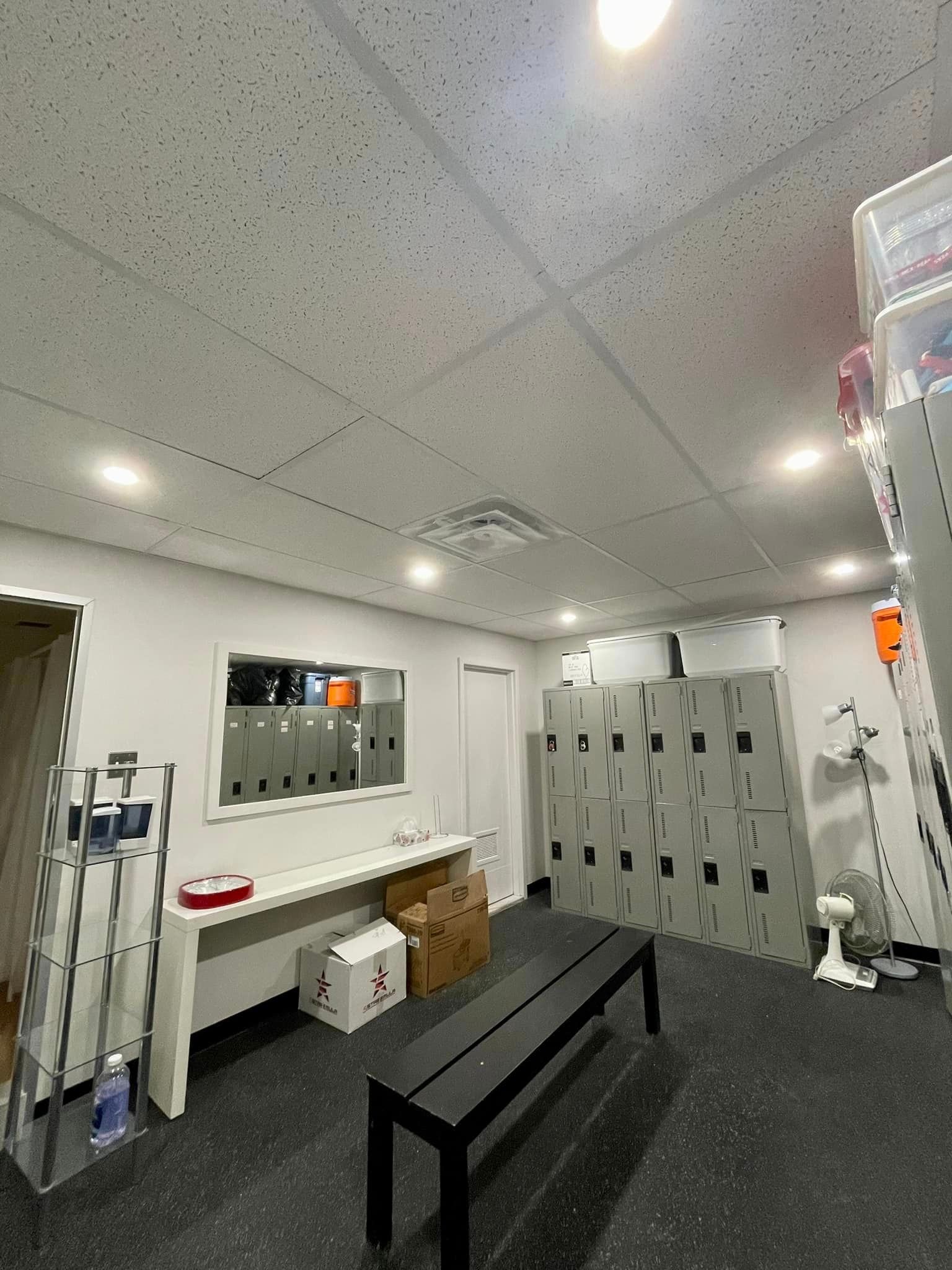 Locker room with black bench, lockers, and a window. Fluorescent lights are on. Gray walls and dark floor.
