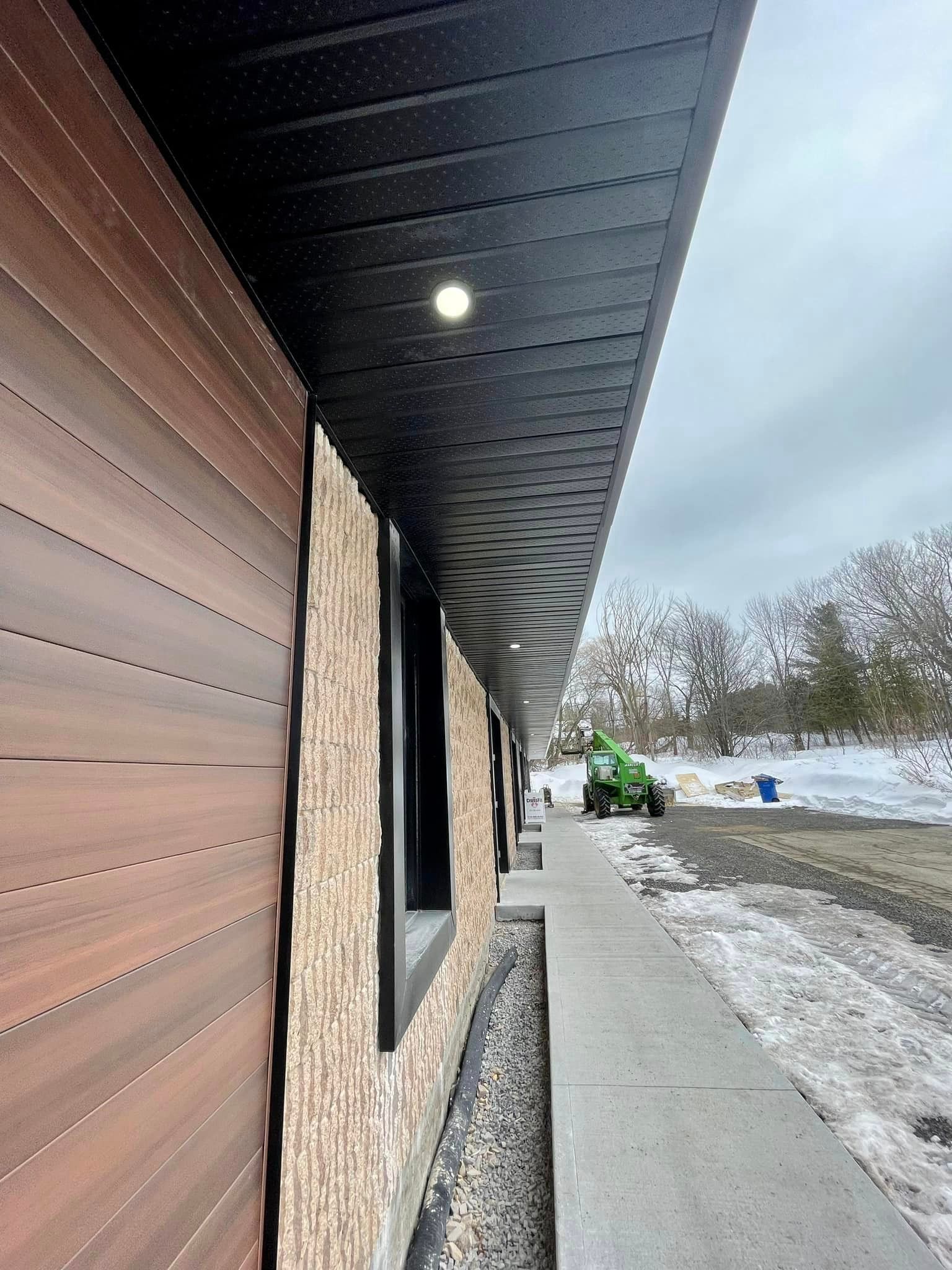 Exterior of a building with brown siding, a black soffit, and a sidewalk. A green machine is in the distance.