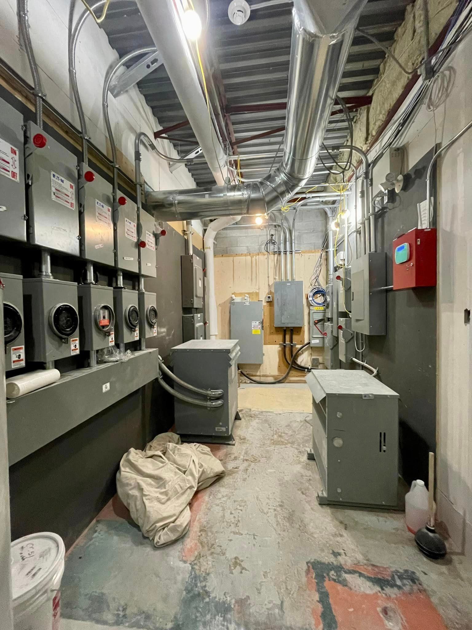 Electrical room with multiple grey panels, wiring, and equipment; concrete floor and exposed ceiling.