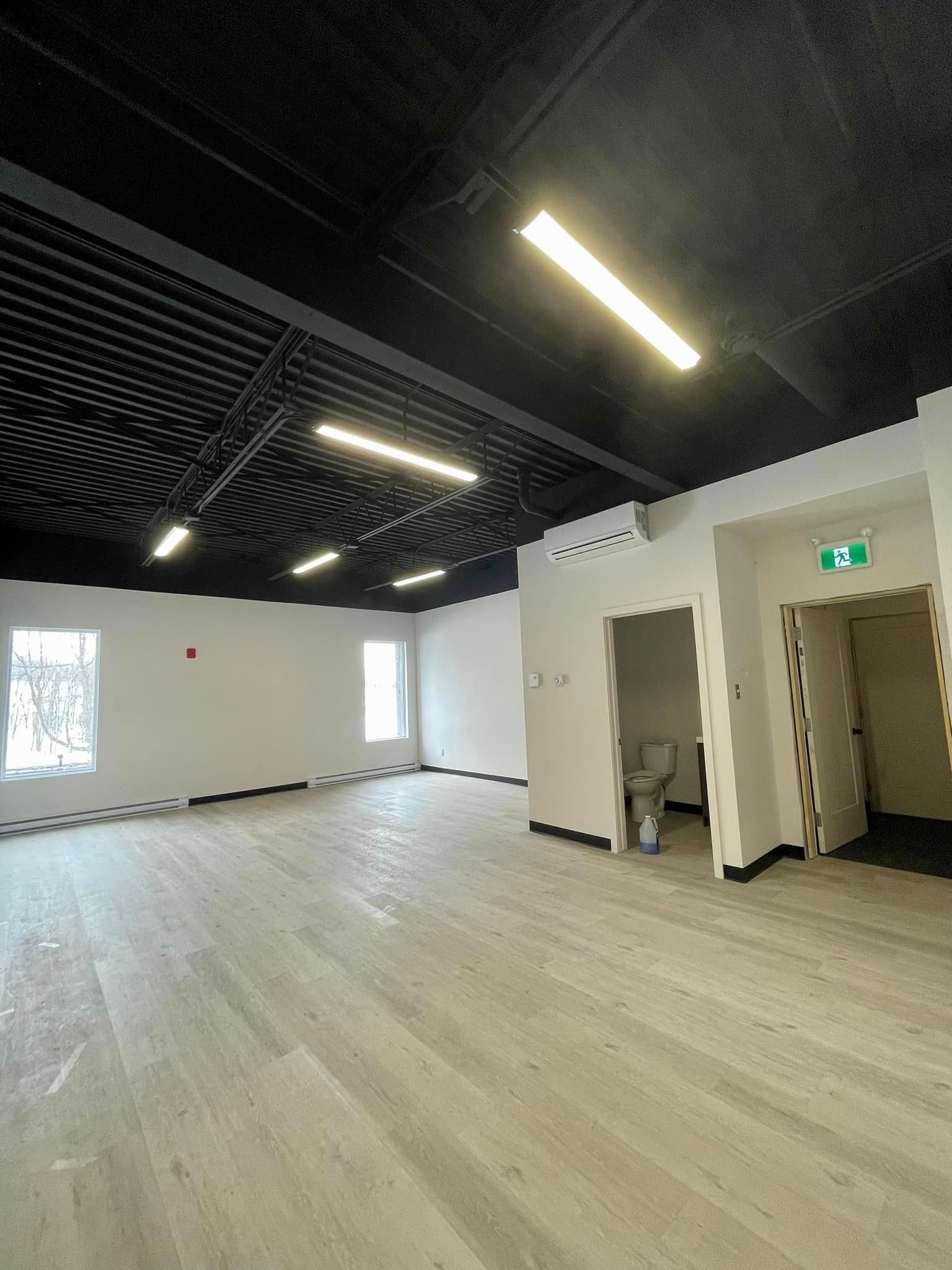 Empty, bright room with gray floors, white walls, and black ceiling with recessed lighting; doorway to a bathroom.