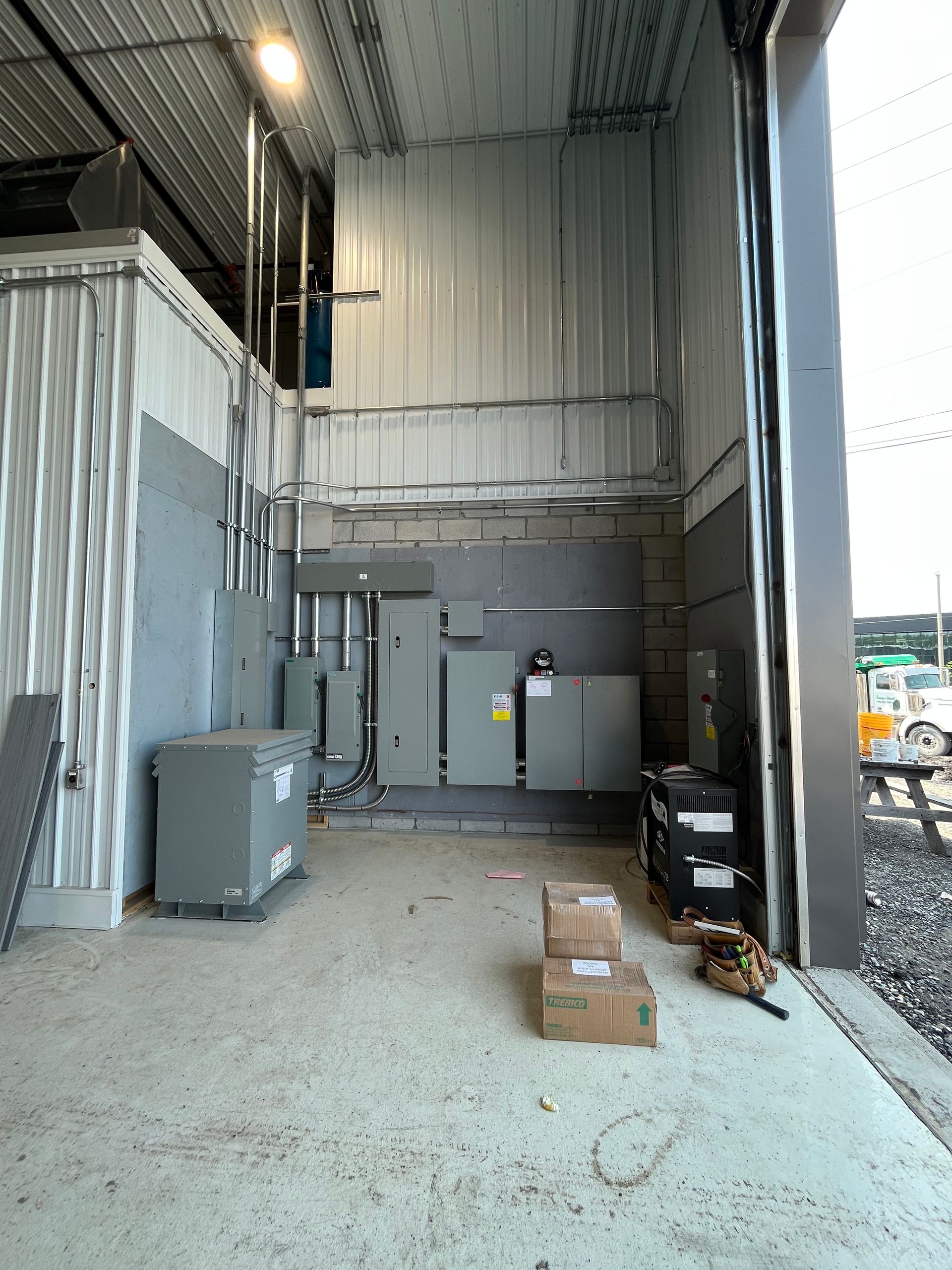 Electrical equipment room with gray transformers and panels, boxes on the floor, and an open garage door.