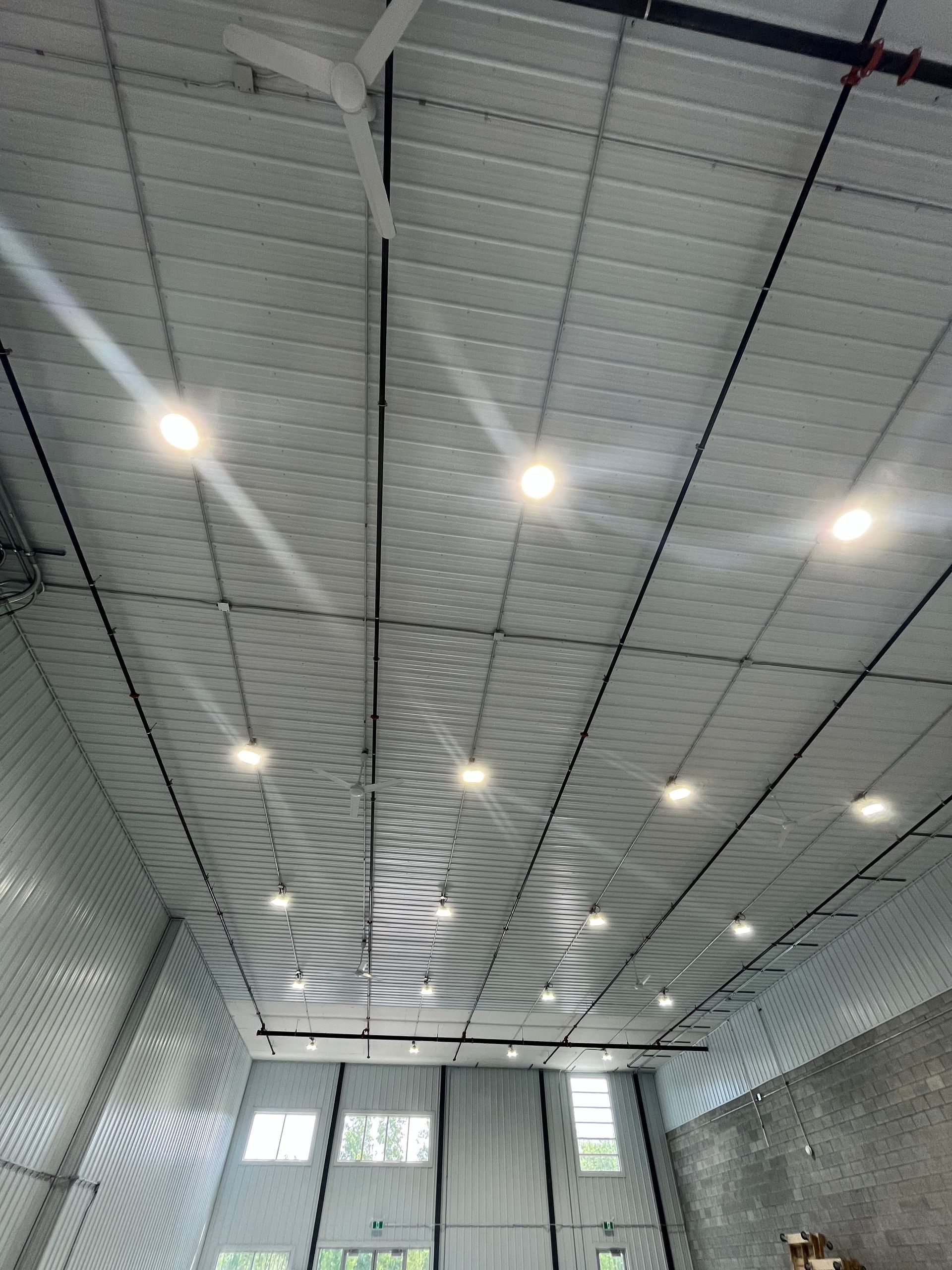Interior view of a building with a high, metal roof, lights, and black wiring. Windows are visible at the bottom.