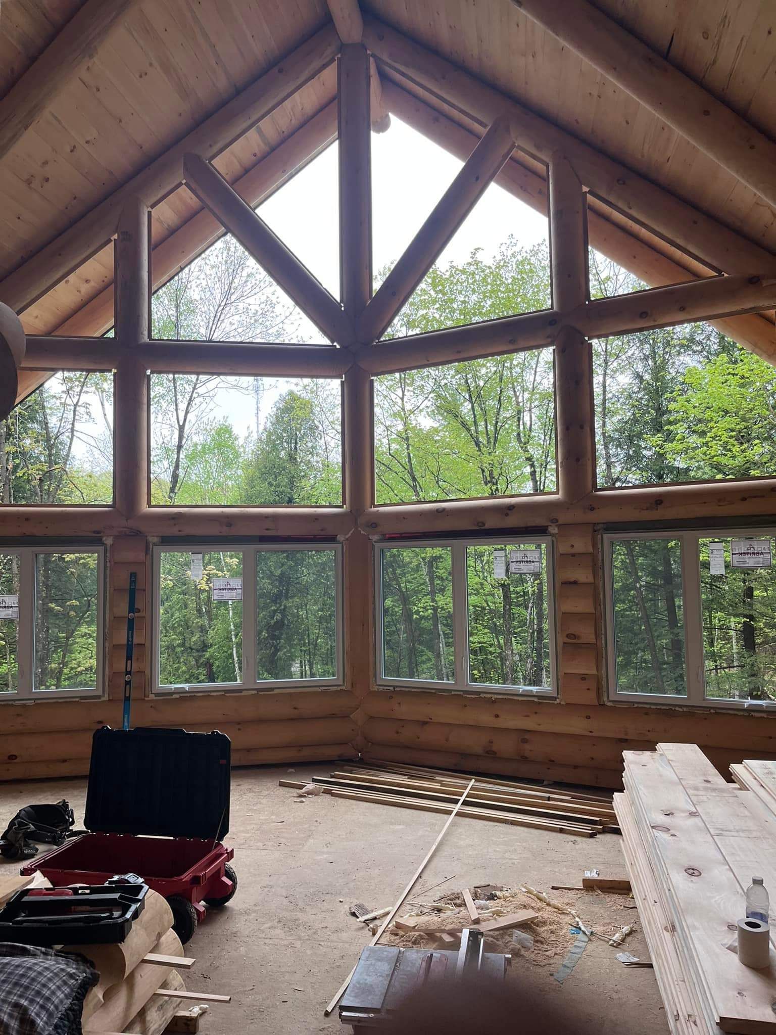 Interior of a log cabin with large windows overlooking trees. Construction tools are on the floor.