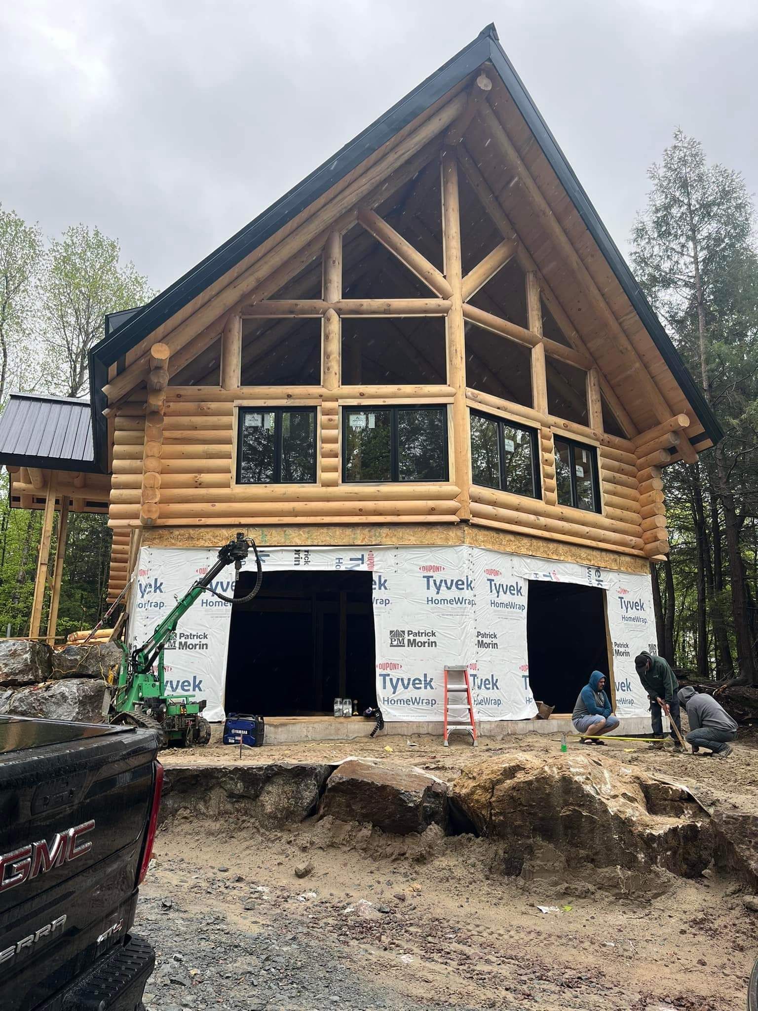 Log cabin under construction with large windows and a high roof. Workers outside.