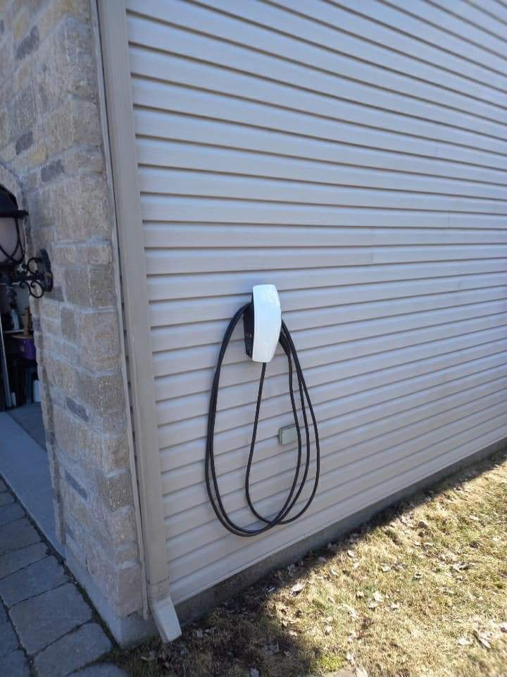 White EV charger mounted on the side of a house with beige siding, black cable coiled below.