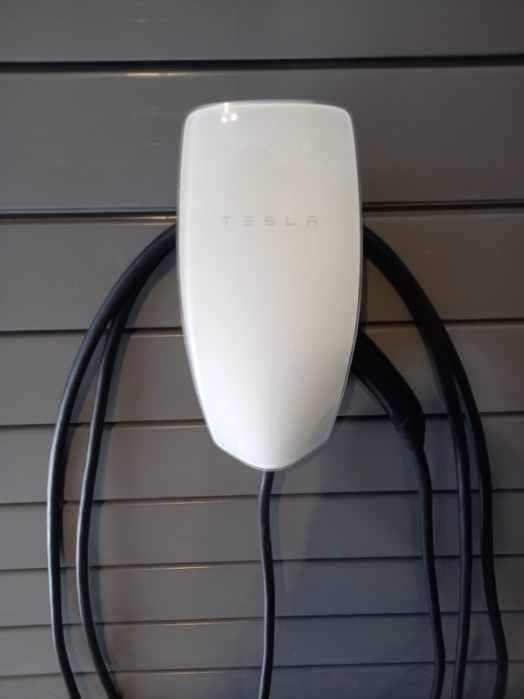 Chargeur mural Tesla blanc avec câble noir, fixé sur un mur gris.
