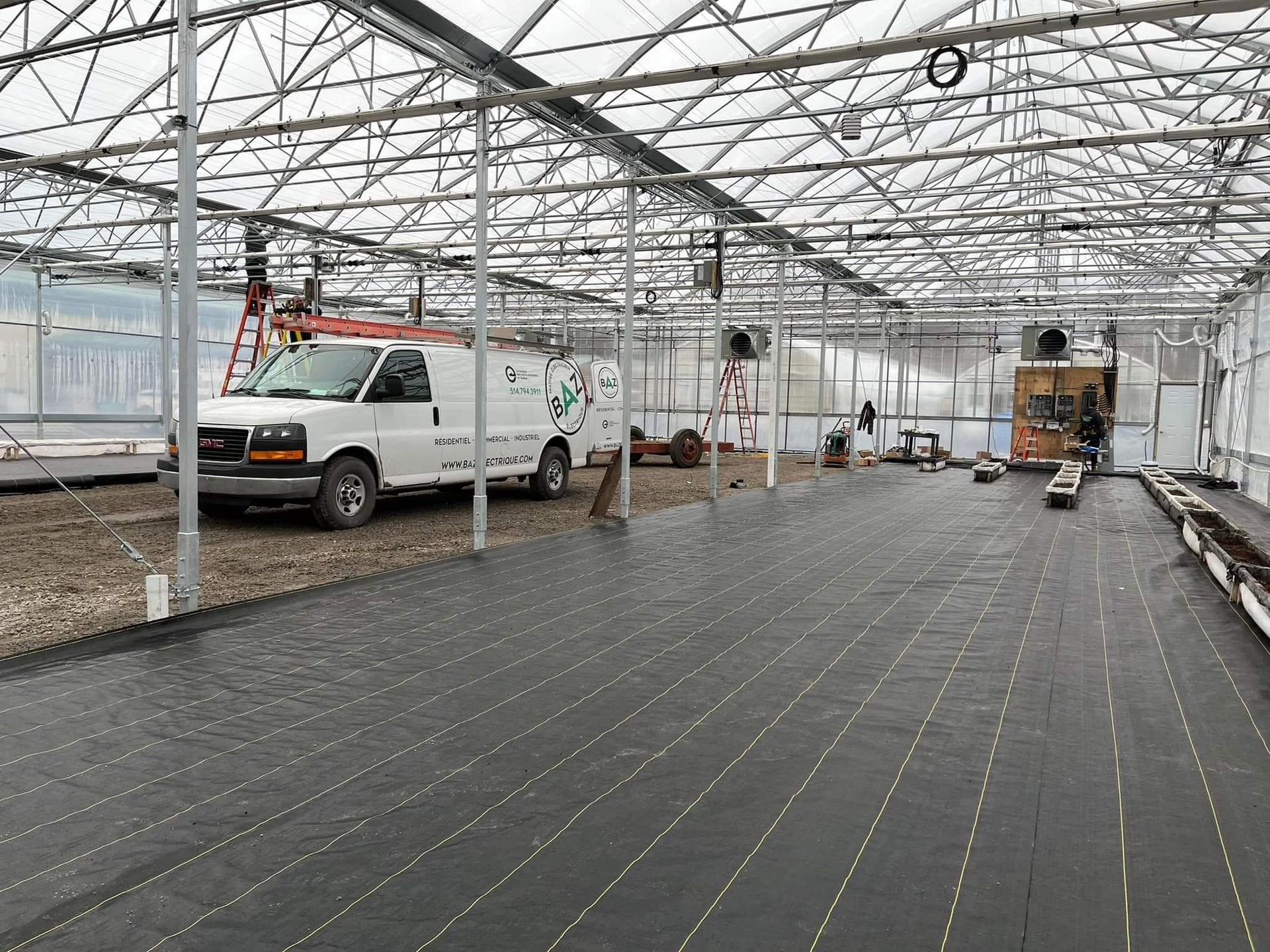 Interior view of a greenhouse with a white van, equipment, and black ground cover.