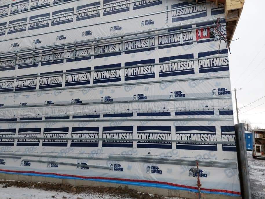Exterior wall of a building under construction, covered in white waterproof membrane, horizontal metal rails.