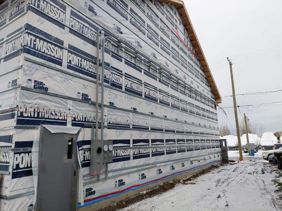 Exterior of a building under construction, wrapped in blue and white material, with electrical conduits and a gray electrical box. Snowy ground.