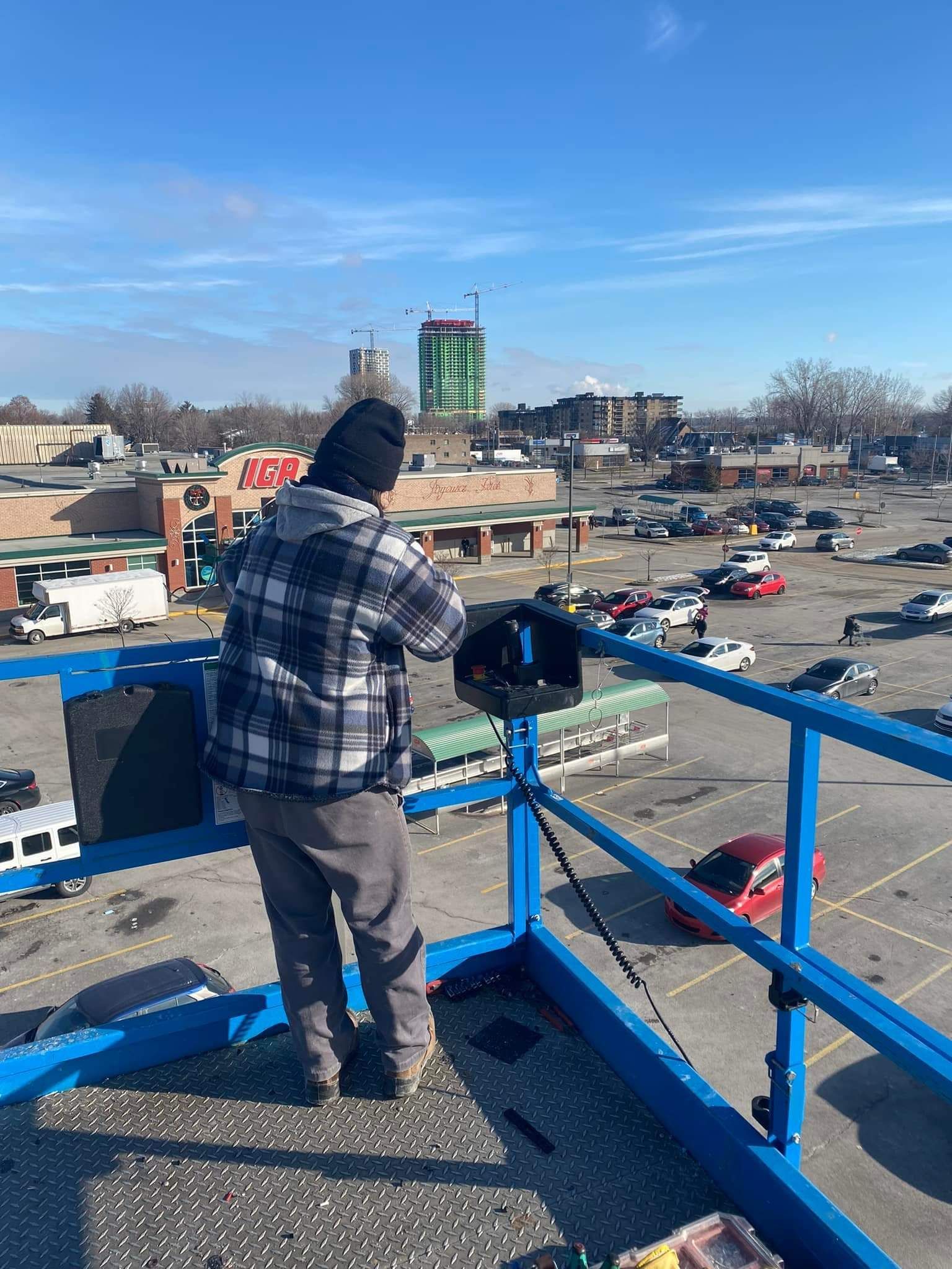 Person on a blue lift platform looking over a parking lot, wearing a plaid shirt and beanie on a sunny day.