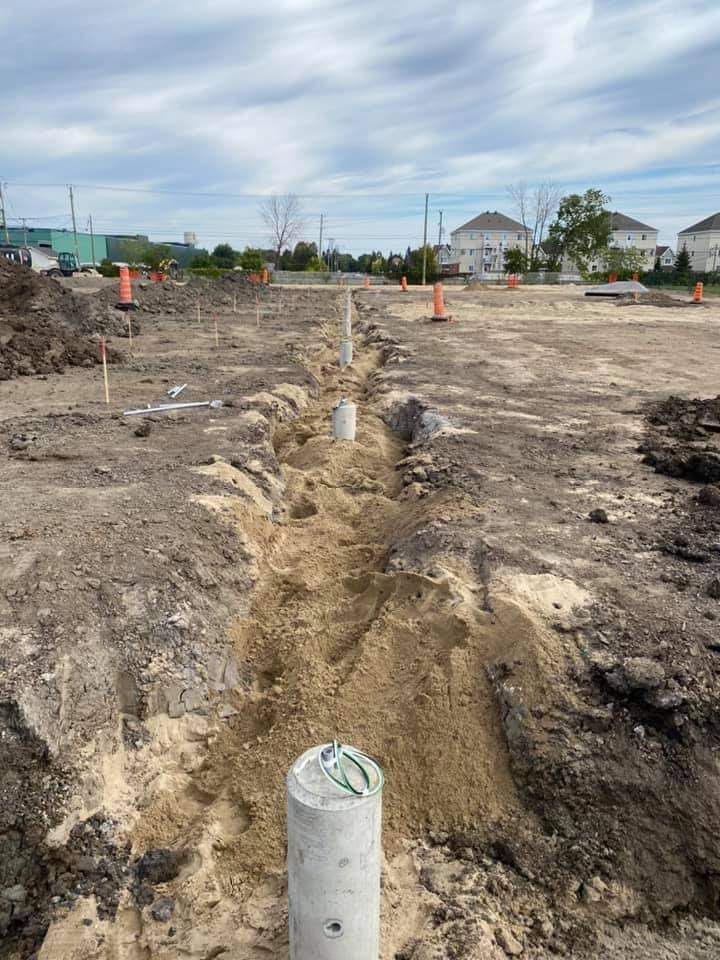 Construction site with a long trench, concrete pillars, and safety cones. Overcast sky.