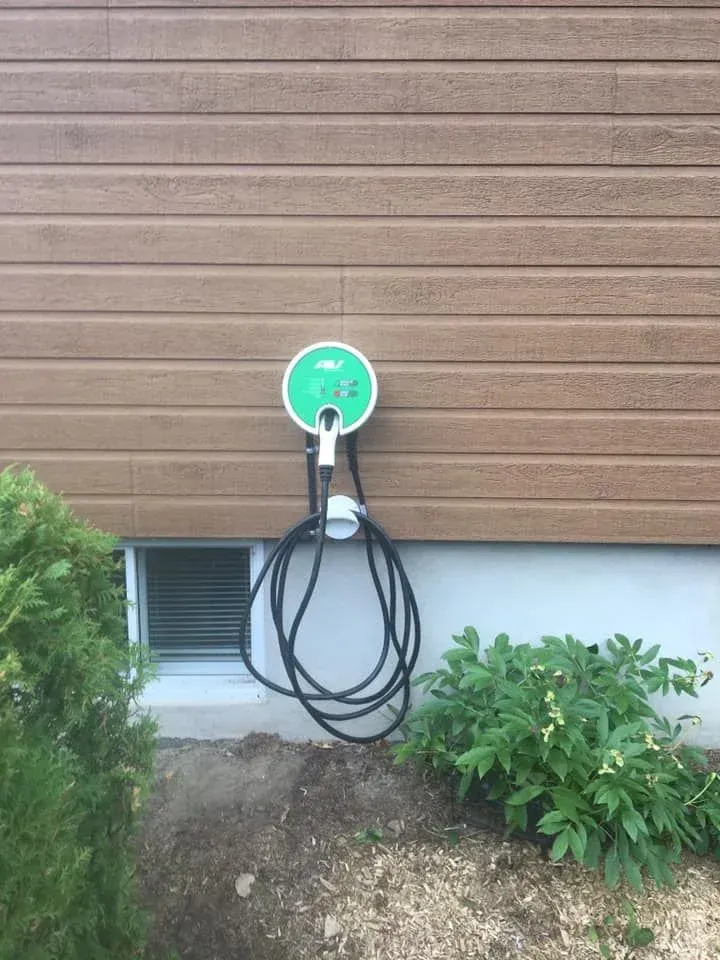 Borne de recharge pour véhicule électrique fixée sur un mur marron, avec un câble noir enroulé en dessous, près d'une fenêtre et de verdure.