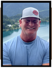 A man wearing a hat and a blue shirt is smiling in front of a lake