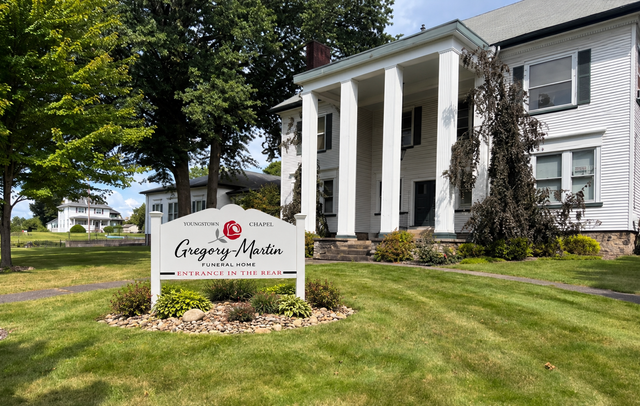 White-columned house with a lawn sign reading Gregory Apria, set among trees and grass.