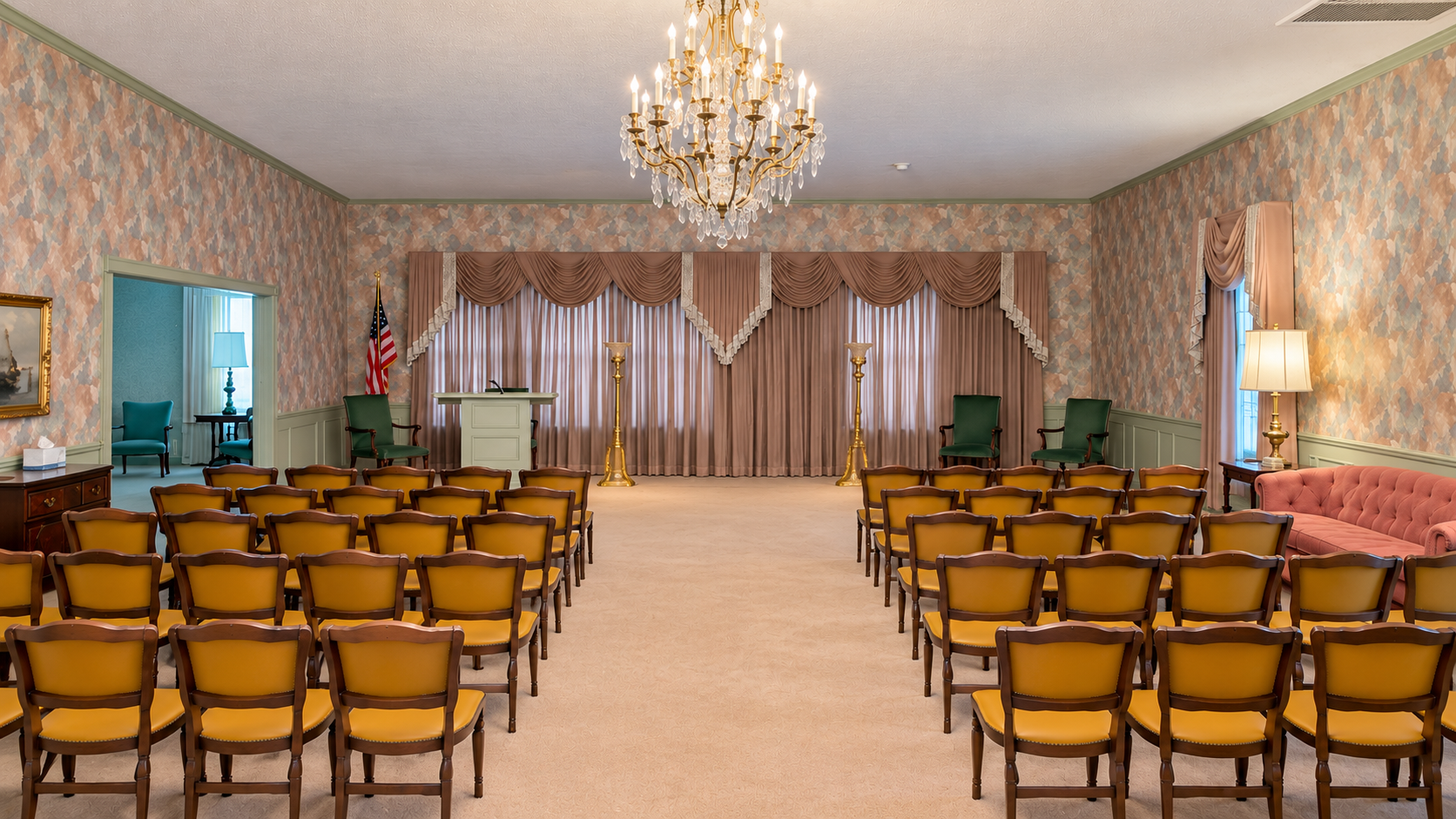 Empty event hall with rows of wooden chairs facing a curtained stage, chandelier overhead.