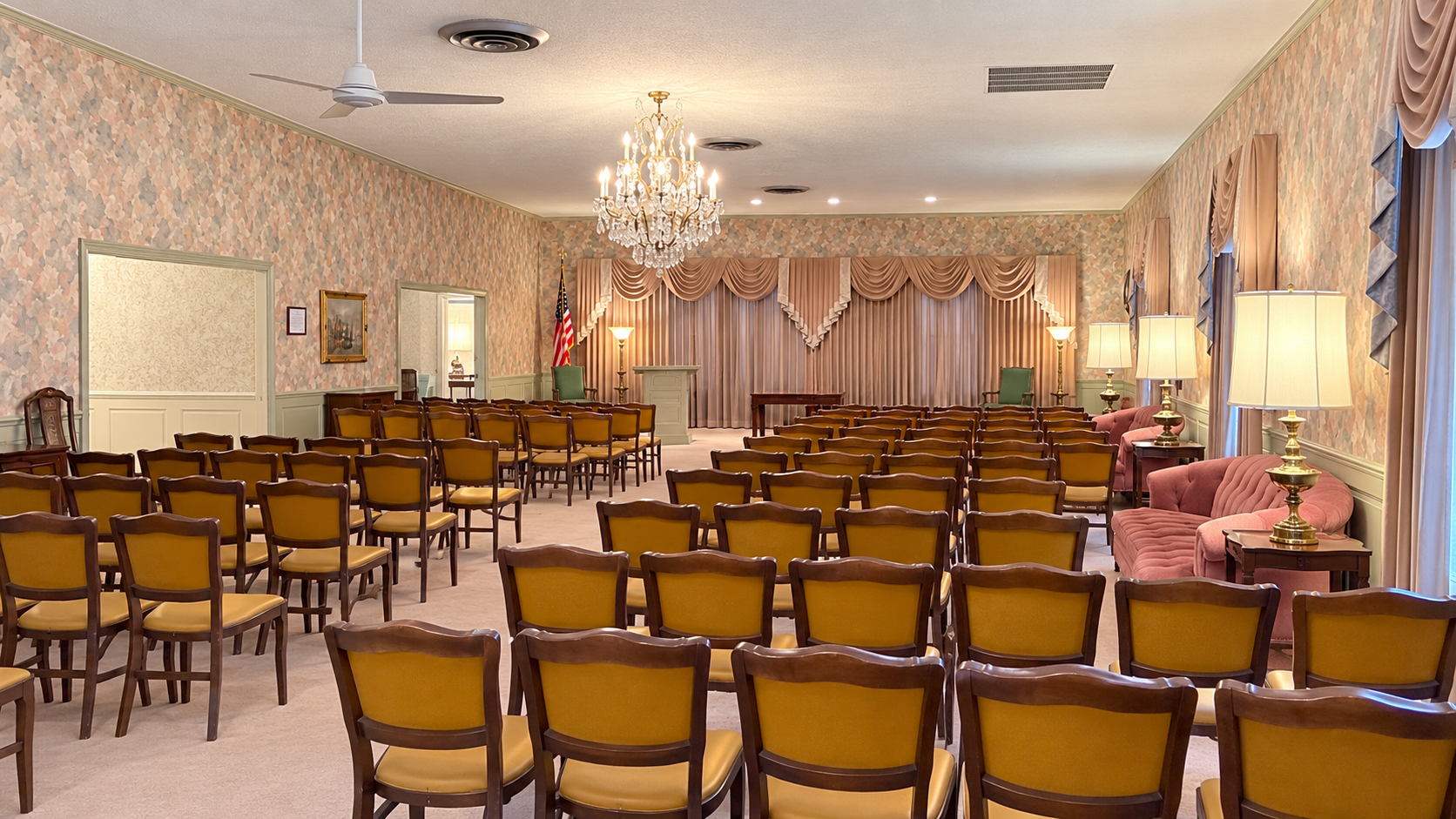 Elegant banquet hall with rows of gold chairs, round tables, chandeliers, and a decorated stage area