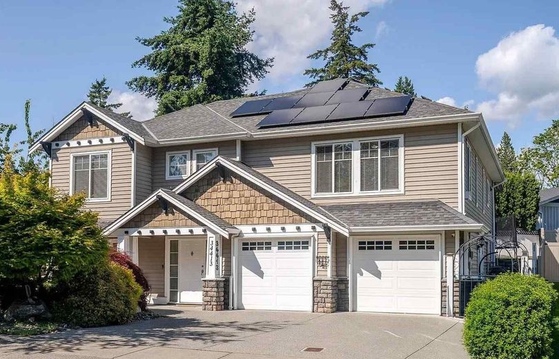 A two-story tan suburban house with solar panels on the roof and an attached two-car garage under a sunny blue sky.