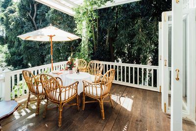 A round table with four wicker chairs on a wooden deck under a white umbrella, surrounded by lush green trees.