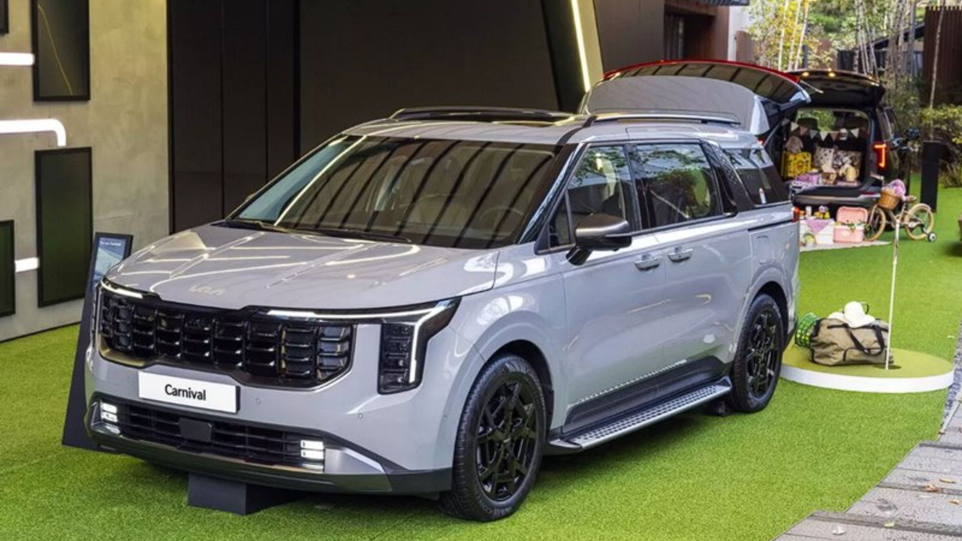 Silver SUV displayed indoors on green turf at an auto show, with luggage on the roof rack