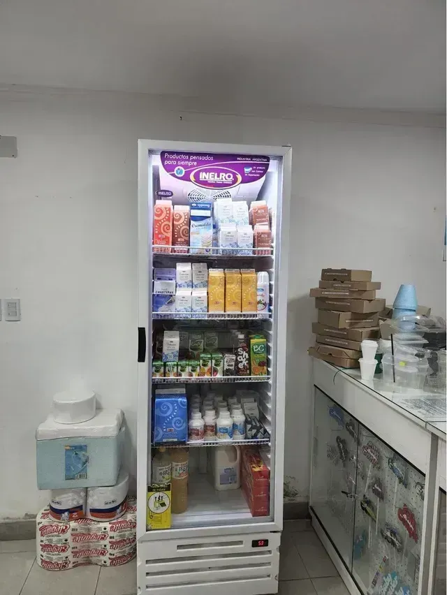 Pequeño refrigerador de tienda con bebidas y tentempiés, junto a un mostrador y un dispensador de agua.