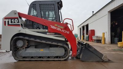 Takeuchi TL10 compact track loader, red and gray, with bucket, parked outside a building.