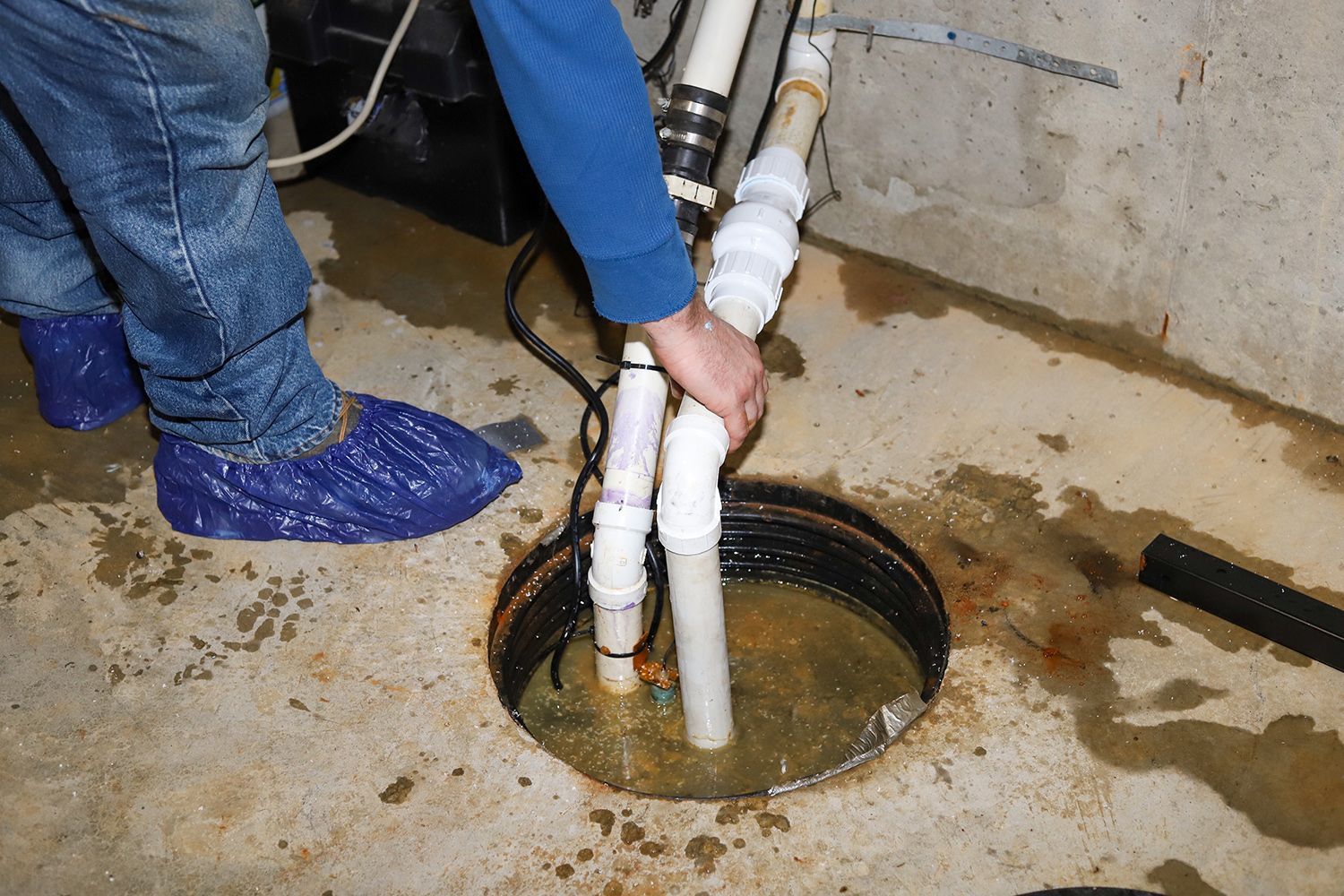 A worker adjusts pipes in a flooded basement sump pump pit. A worker adjusts pipes in a flooded basement sump pump pit.