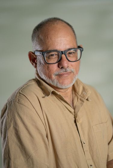 Man in black polo shirt and glasses standing against a black background, smiling with hands clasped.