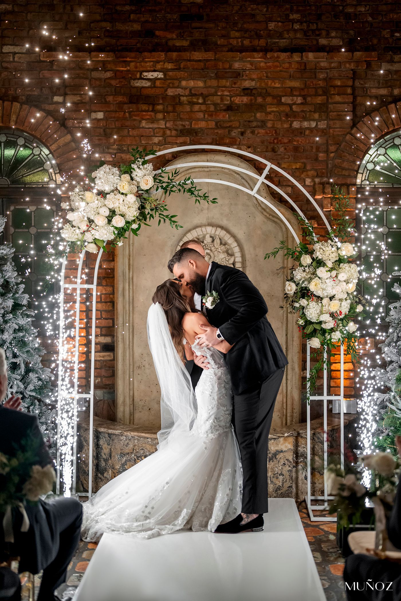 Wedding couple kissing beneath a white floral arch with fairy lights in a brick-walled venue