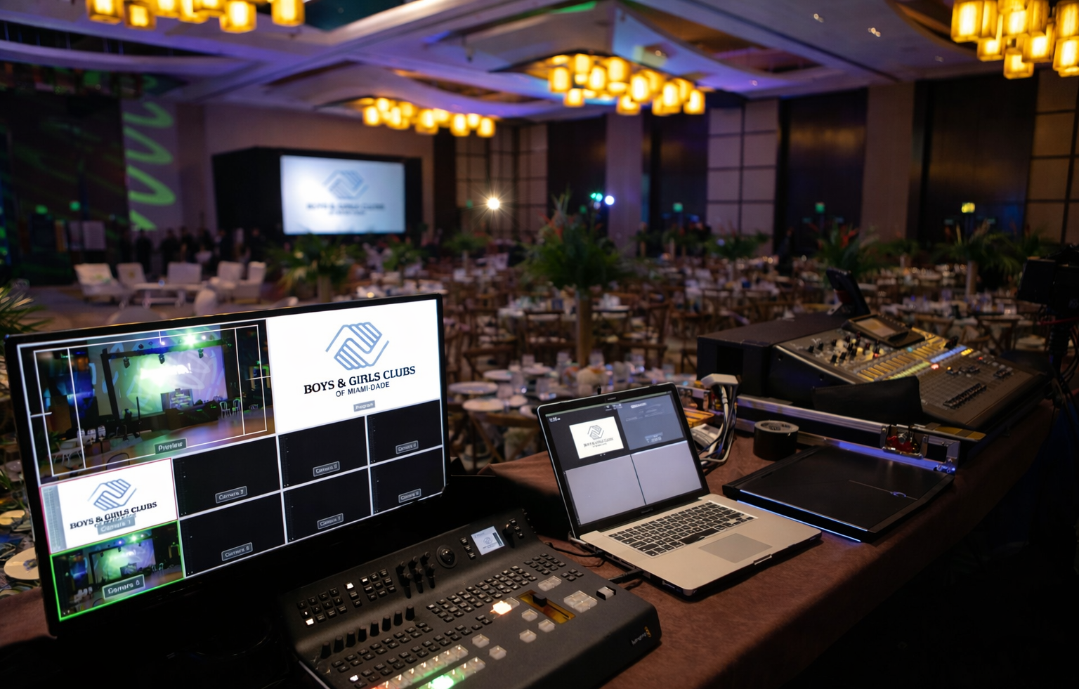 Audio/video control booth at a banquet hall with monitors, laptop, and lighting console overseeing a stage